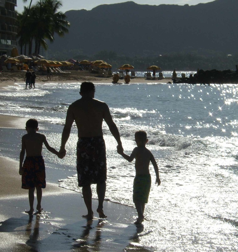 A silhouette of a father holding the hands of two young boys as they walk along the beach